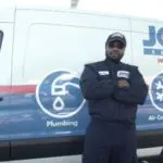 A person in a uniform stands with arms crossed in front of a service van labeled with plumbing and air conditioning logos.