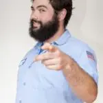 A bearded man in a light blue shirt points forward with a finger while slightly smiling.