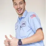 A person wearing a light blue work shirt with patches and an American flag on the sleeve is smiling and giving a thumbs-up gesture.