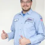 A person in a blue uniform and cap is smiling and giving two thumbs up. The uniform has a name patch and an American flag patch.