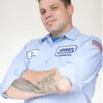 Person in a light blue work shirt with company logo, arms crossed. Visible tattoos on forearm and an American flag patch on the sleeve.