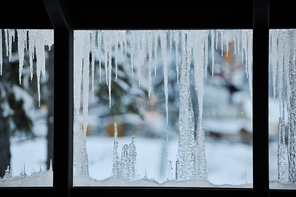 Icicles hang from the edge of a roof, with snow-covered trees and a blurred background visible through the window.