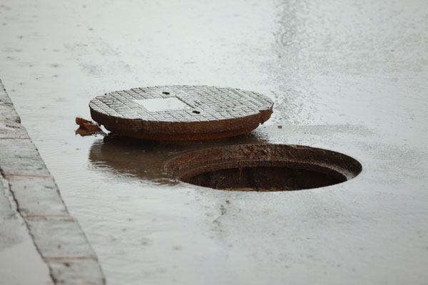 A partially open manhole cover on a wet street, with water pooling around it.