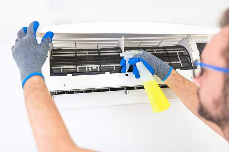 Person cleaning an air conditioning unit with a spray bottle, wearing gloves and safety glasses.