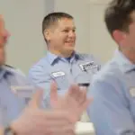 Three uniformed individuals are clapping in a bright room, with one person in focus and smiling.