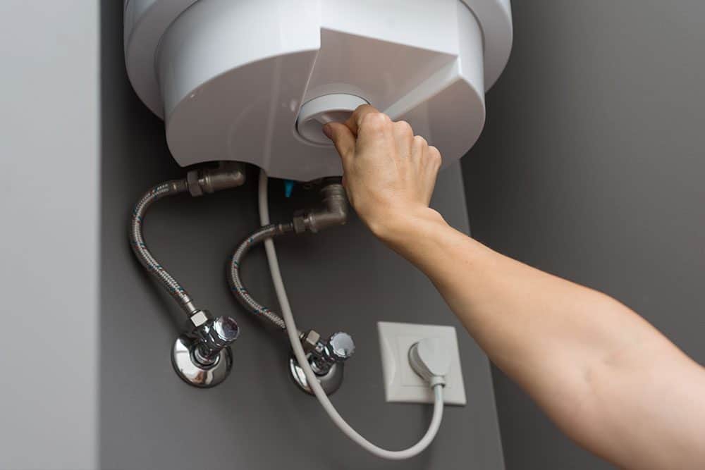 A person adjusts a dial on a wall-mounted water heater with connected pipes and a visible power outlet.
