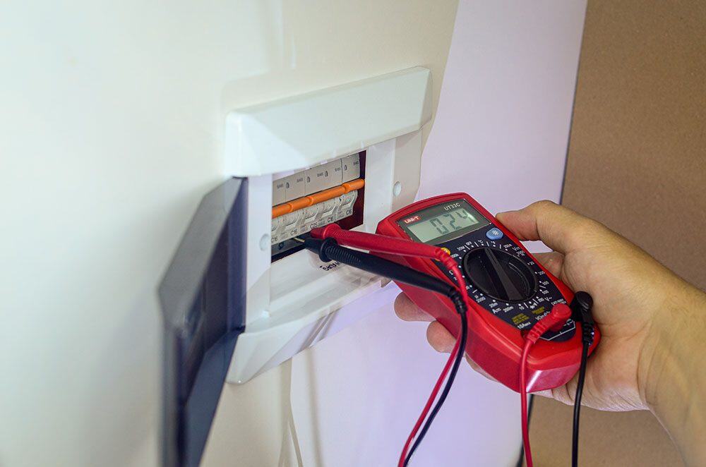 A person uses a red digital multimeter to check voltage in a circuit breaker box.