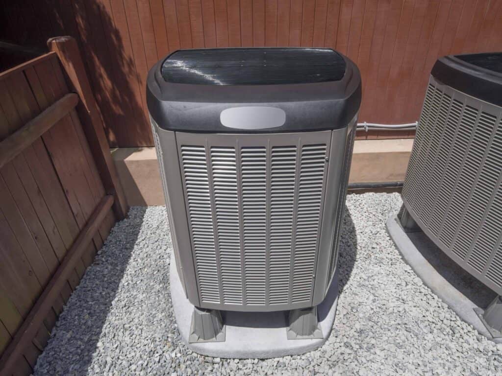 Outdoor air conditioning unit placed on a gravel bed next to a wooden fence and wall.