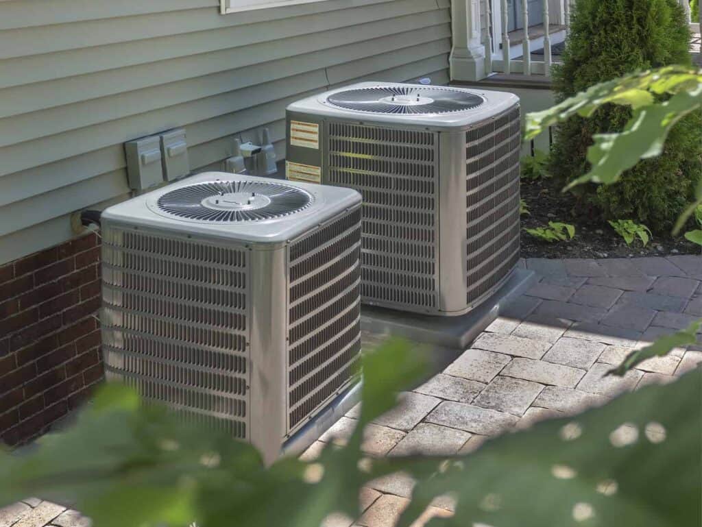 Two outdoor air conditioning units sit on a paved area beside a building with siding and a brick wall.