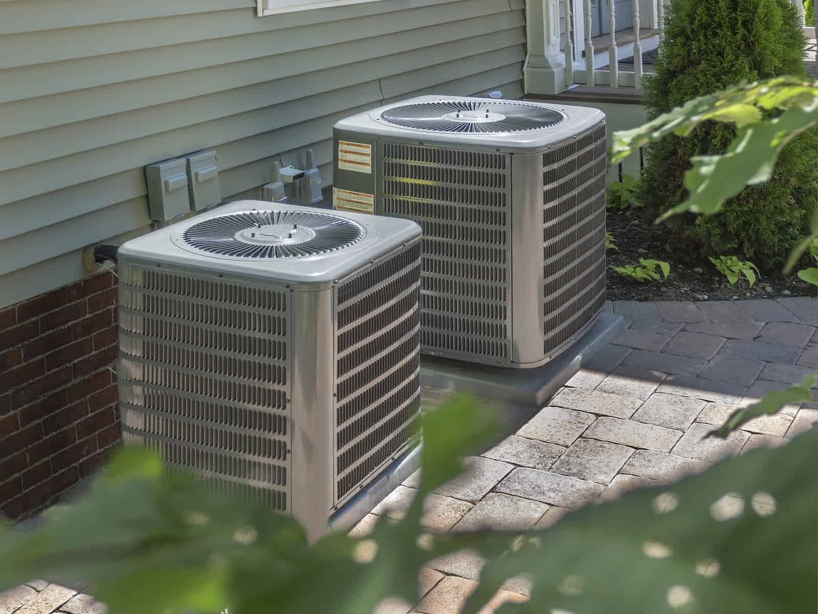 Two outdoor air conditioning units sit on a paved area beside a building with siding and a brick wall.