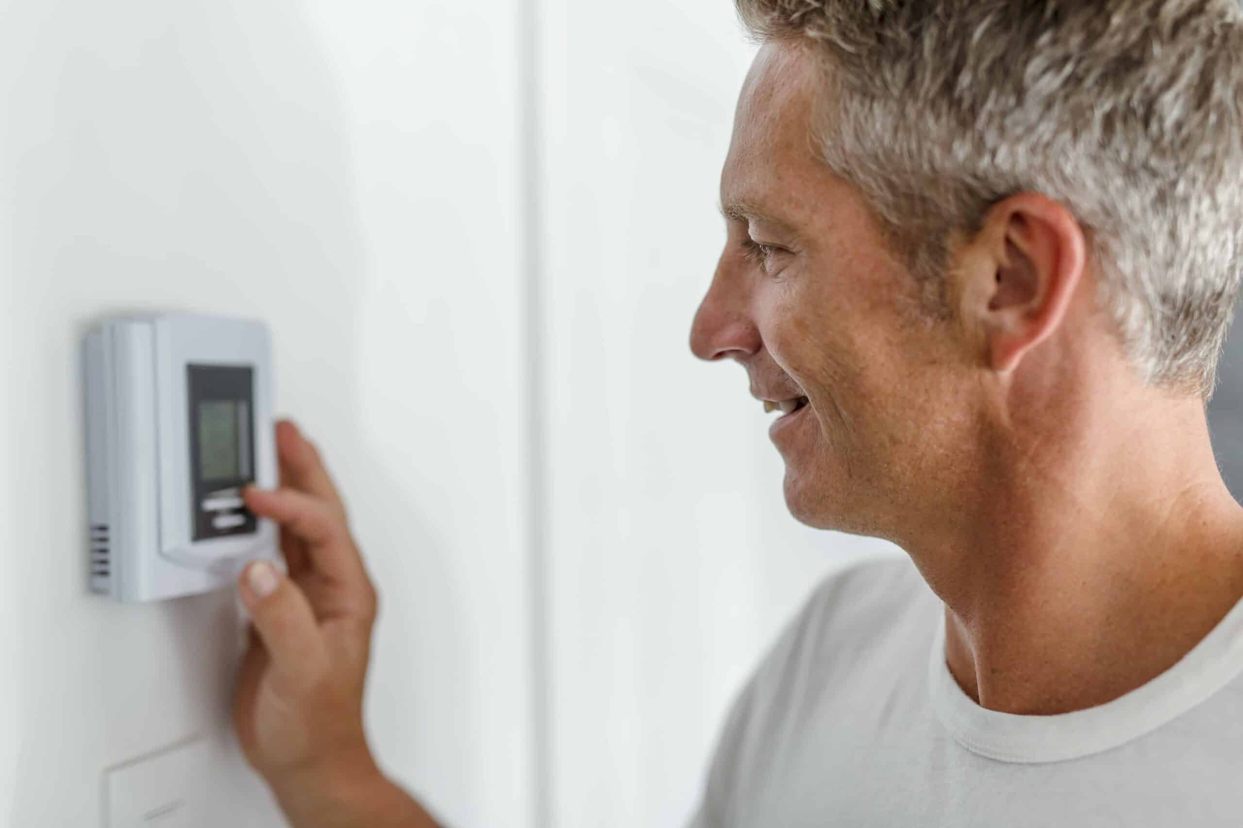 Person adjusting a wall-mounted digital thermostat, smiling while pressing buttons, wearing a white shirt.