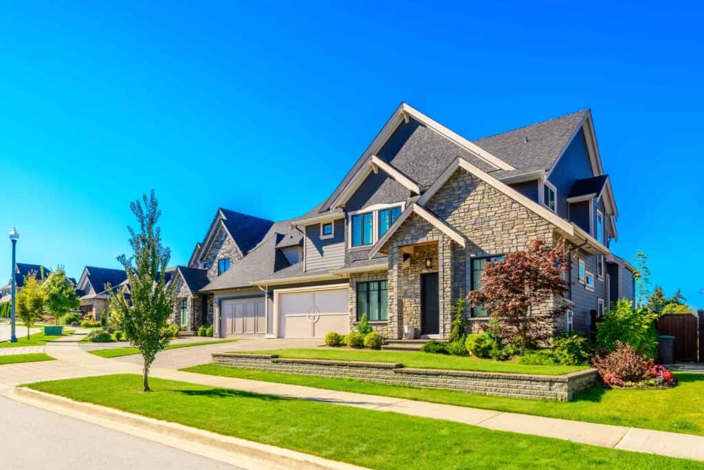 A row of modern suburban houses with stone and siding exteriors, neatly manicured lawns, and trees, under a clear blue sky.