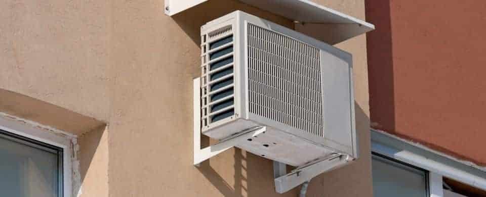 Outdoor air conditioning unit mounted on a beige wall between two windows.