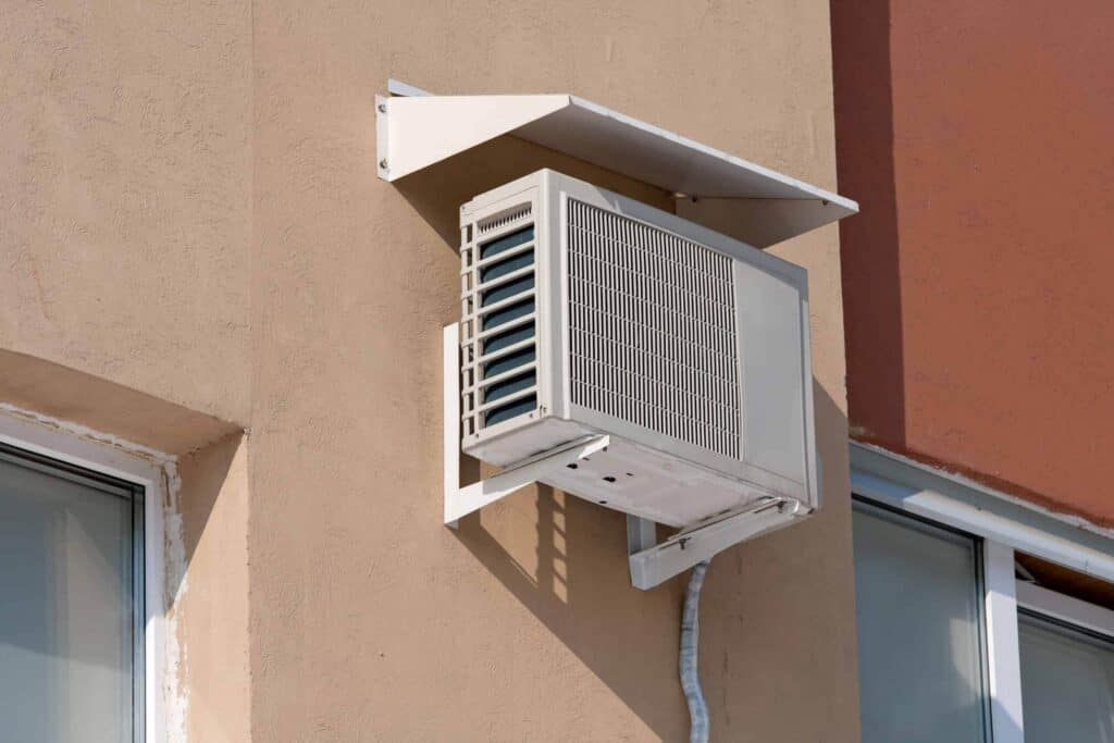 Wall-mounted air conditioning unit outside a building, situated between two windows, with a protective cover on top.