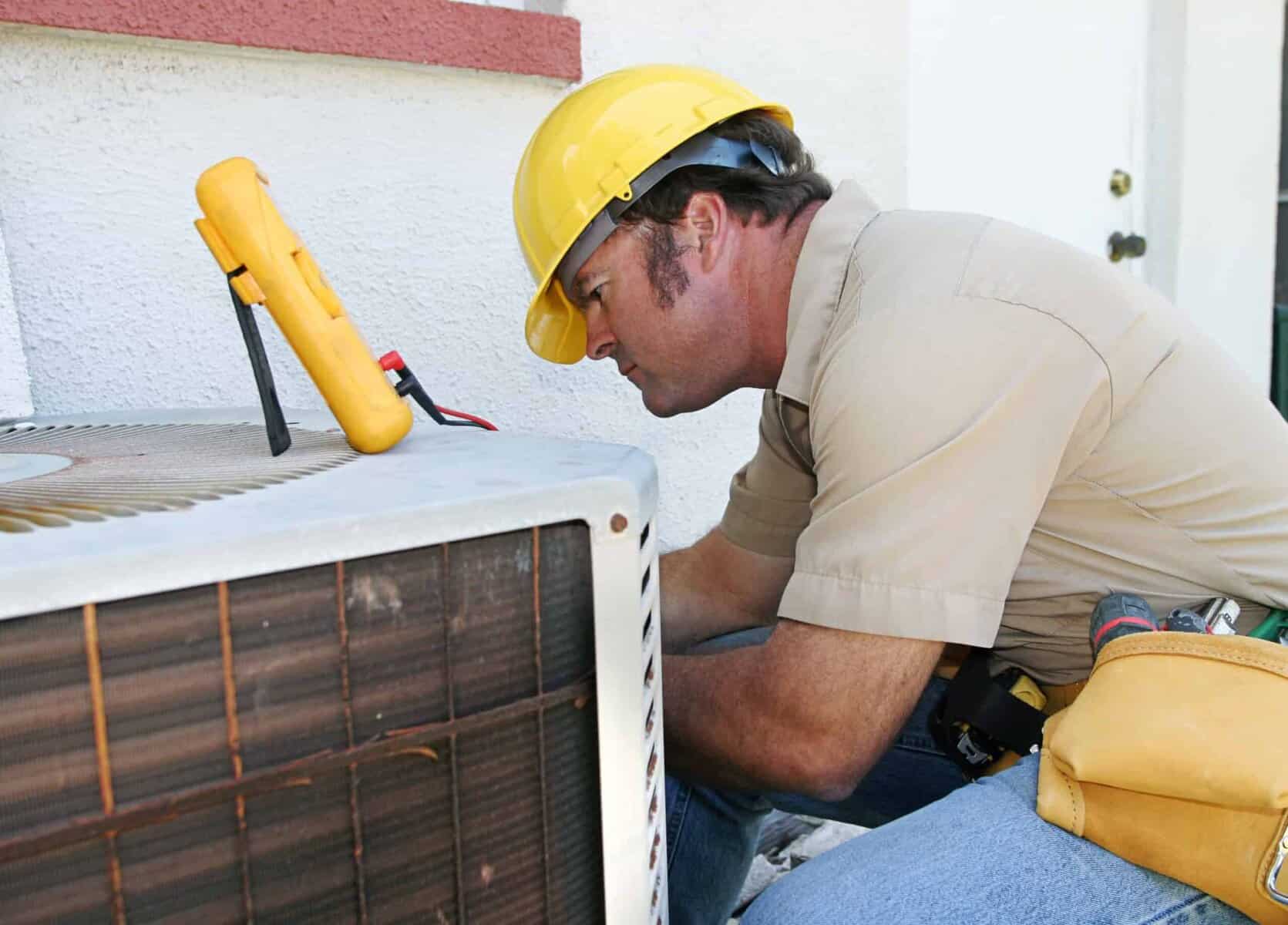 Technician in a yellow hard hat inspects an outdoor air conditioning unit using a multimeter.