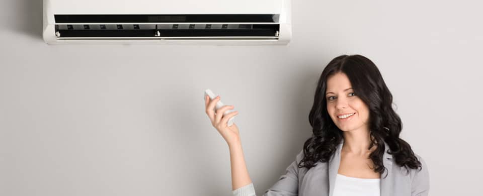A woman with long dark hair holds a remote control and smiles while standing near a wall-mounted air conditioning unit.