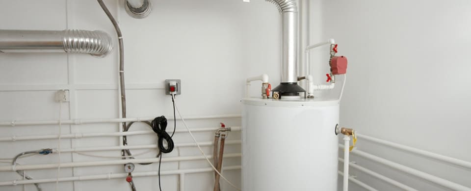 A white water heater is installed in a room with white walls and multiple pipes. A plug is connected to an electrical outlet.