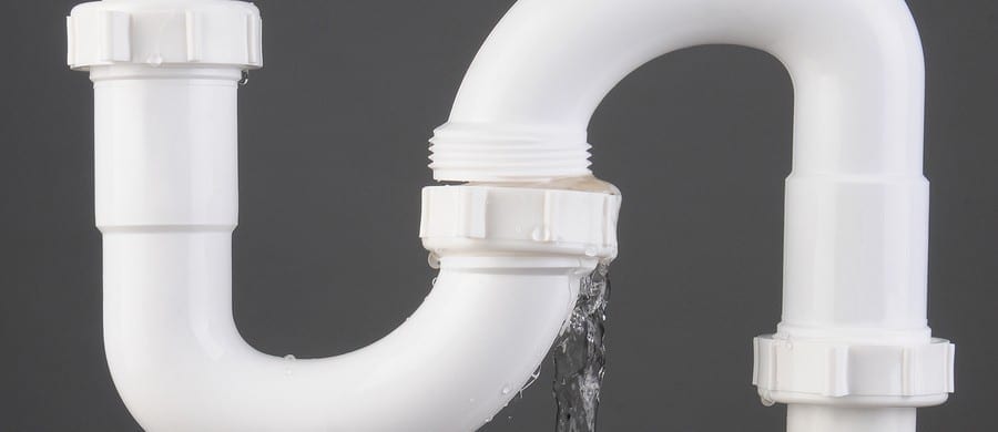 Close-up of a leaking white plastic pipe with water dripping at a joint against a gray background.