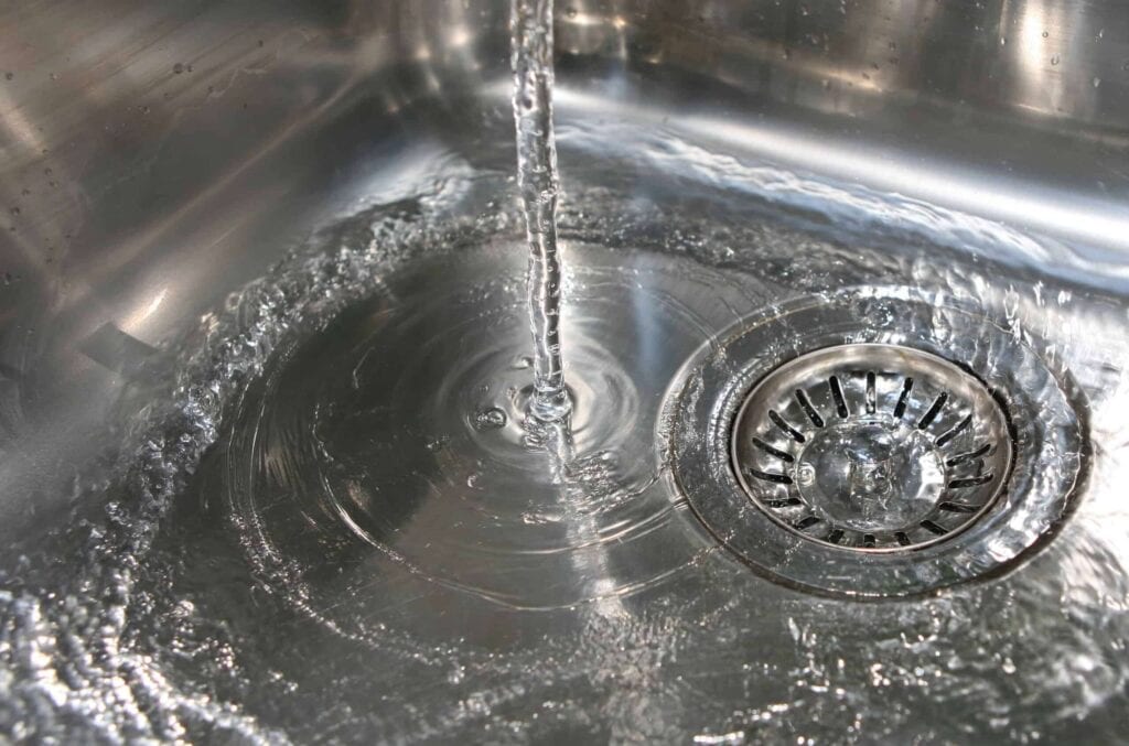 Water flowing from a faucet into a stainless steel sink with visible drain and swirling water.