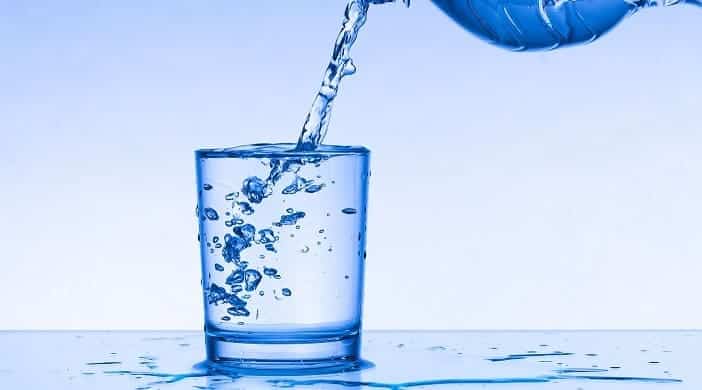 Water is being poured from a container into a transparent glass with water splashing out, on a smooth blue surface against a blue and white gradient background.