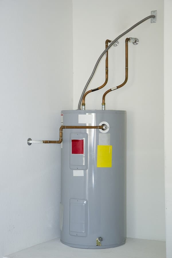 A gray water heater installed in a corner with exposed copper and steel pipes connecting at the top and side, against a white wall.