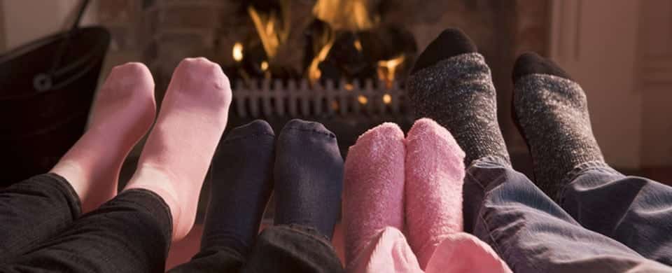 Four pairs of feet, wearing socks, are stretched out in front of a fireplace.