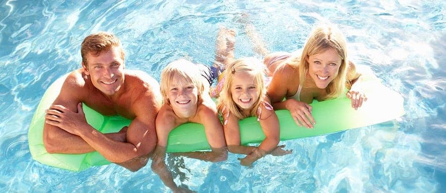 Four people smiling while leaning on a green pool float in clear blue water.