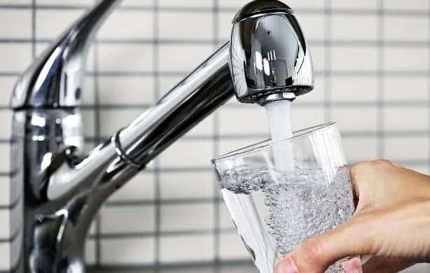 A person fills a clear glass with water from a shiny chrome faucet against a background of white tiled wall.
