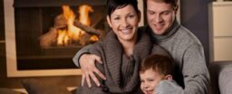 A family of three sits together in front of a fireplace. The parents wear gray sweaters, and the child is in front, smiling. The room is warmly lit, creating a cozy atmosphere.