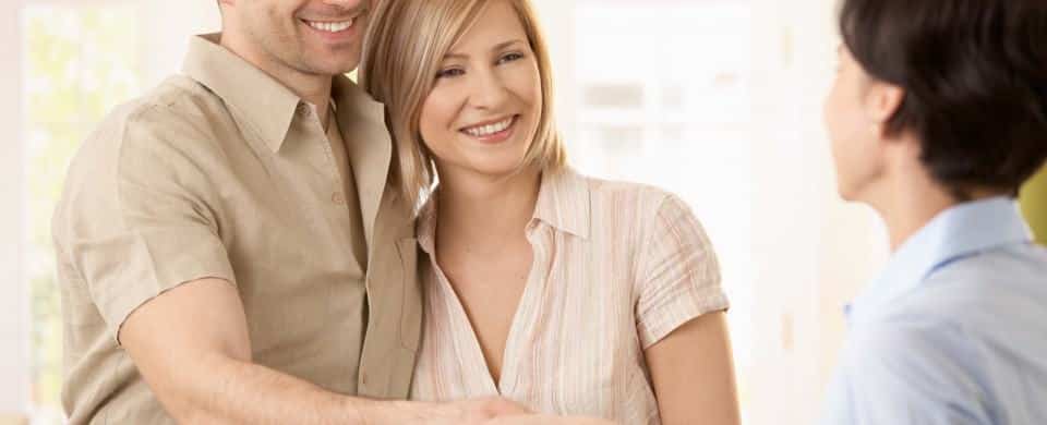 A smiling couple stands together while greeting and shaking hands with a Jones Services Plumbing Contractor indoors.