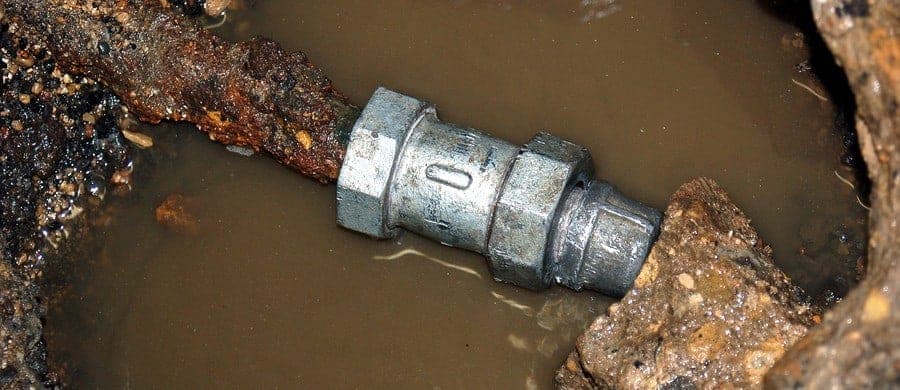 A damaged rusty pipe with a metal connector is partially submerged in muddy water, showing signs of corrosion and decay.