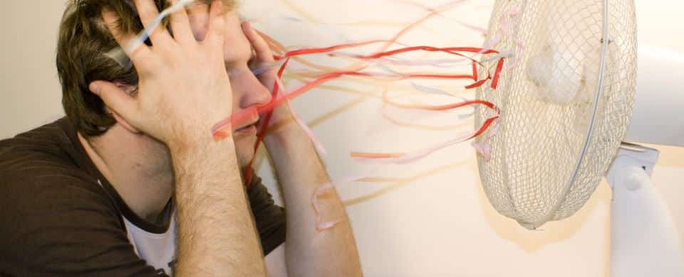 Person with hands on head sits facing a fan blowing colorful streamers.