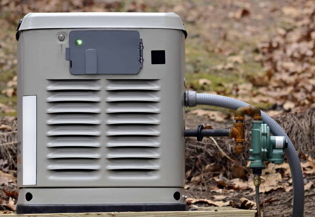 A gray backup generator connected by pipes sits on a wooden platform in a yard with dry leaves scattered on the ground.