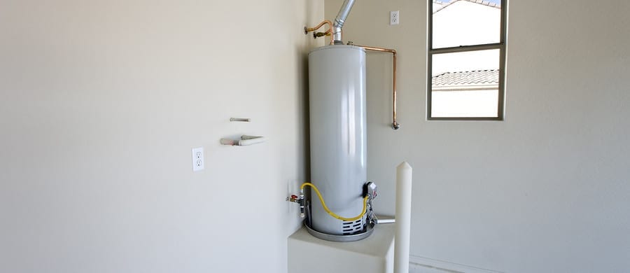 A tall, cylindrical water heater with pipes is installed on a platform against a beige wall in a room with a window.