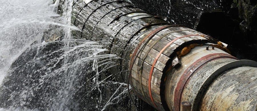 Water gushes from a large, corroded industrial pipe with visible metal bands, indicating signs of wear and leakage.