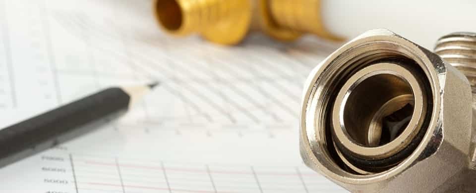 Close-up of a metal plumbing fitting and a pencil on top of a graph paper or technical drawing, with a blurred brass fitting in the background.