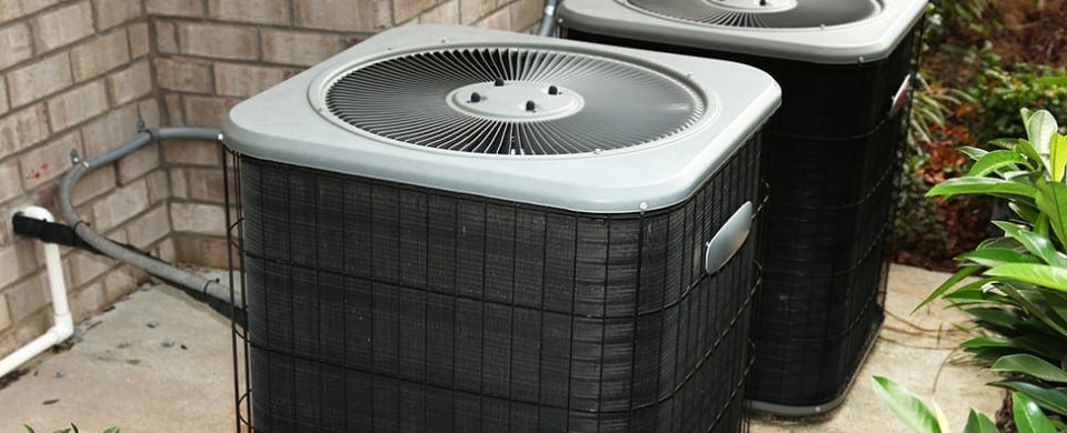 Two outdoor air conditioning units placed on a concrete surface beside a brick wall, surrounded by green shrubs.