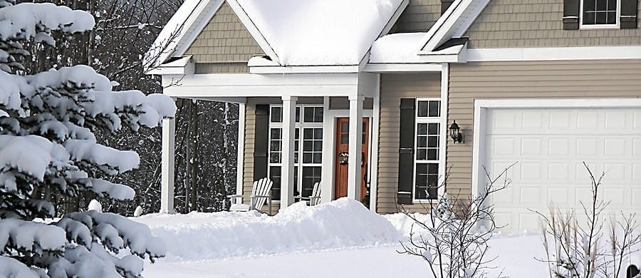 A snow-covered suburban house with a front porch, two chairs, and a large garage. Snow blankets the roof and yard, and evergreen trees stand in the foreground.