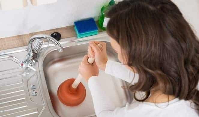 Person using a plunger to unclog a kitchen sink, with a blue sponge and green bottle nearby.