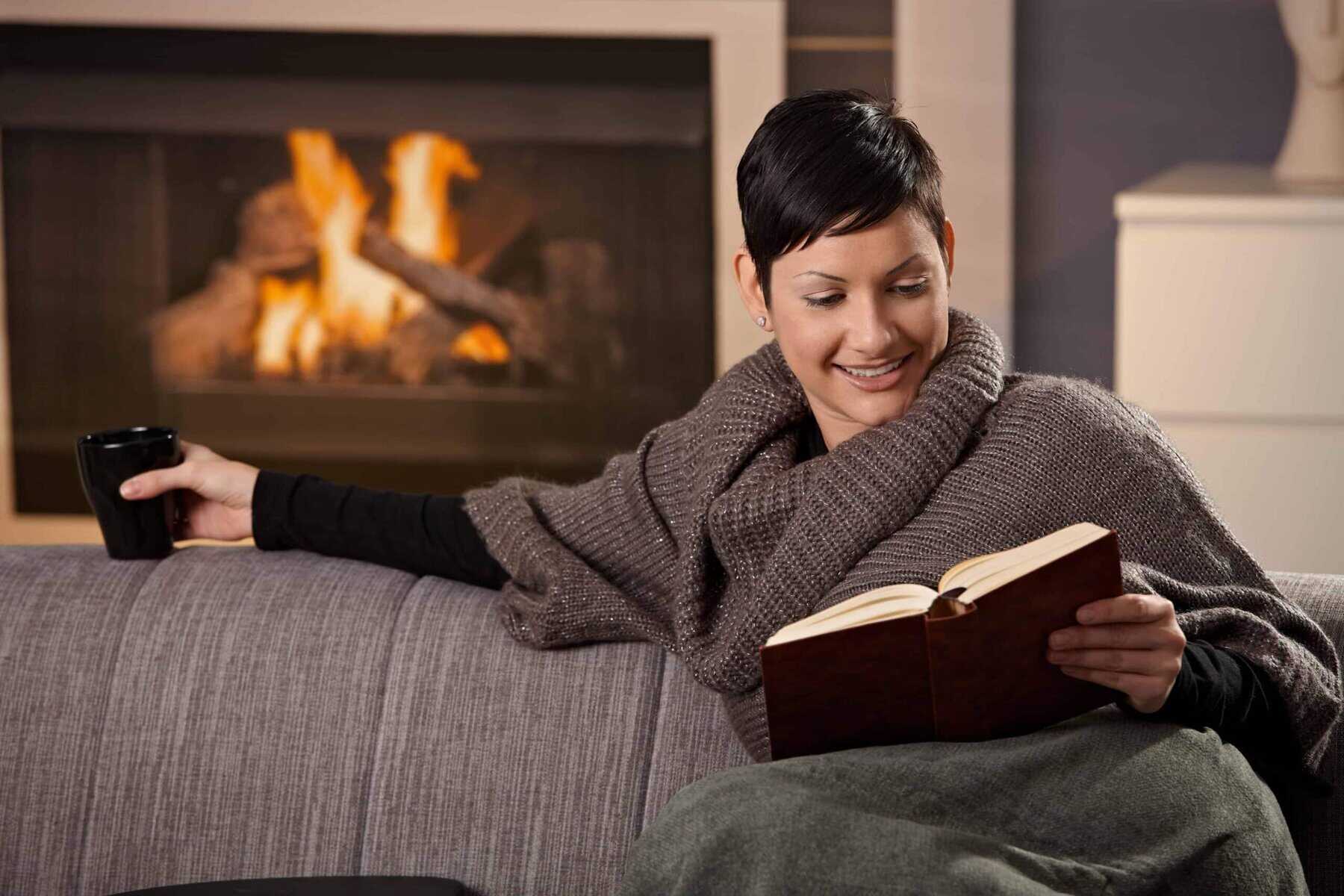 Person with short hair sits on a sofa, wrapped in a blanket and a shawl, holding a book and a mug. A fireplace with burning logs is in the background.
