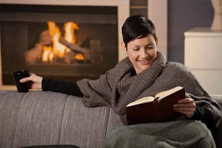 A person sits on a couch covered with a cozy blanket, holding a mug and reading a book, while the warm glow from the fireplace offers comfort akin to glowing service reviews.