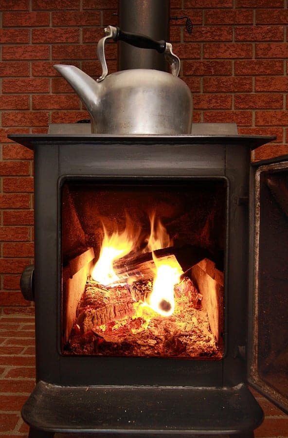 A metal kettle sits on a burning wood stove, with flames visible through the glass door. The stove is placed against a brick wall.