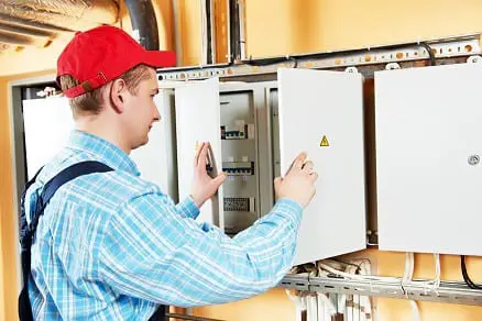 A person in a red cap and plaid shirt meticulously inspects an electrical panel, doors wide open to reveal switches and wiring, ensuring every detail meets the highest standards, echoing the positive service reviews this expert consistently receives.