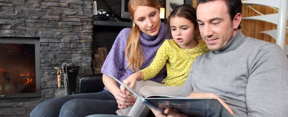 A man and woman sit with a child on a sofa, reading a book together. The background features a stone fireplace with a small fire burning inside.