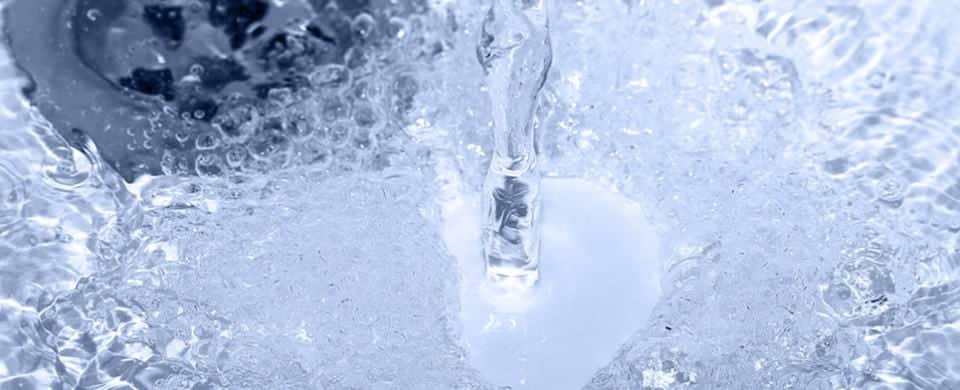 A stream of water flows forcefully from a faucet into a sink, creating splashes and bubbles on the surface below. The sink's drain is partially visible in the background.