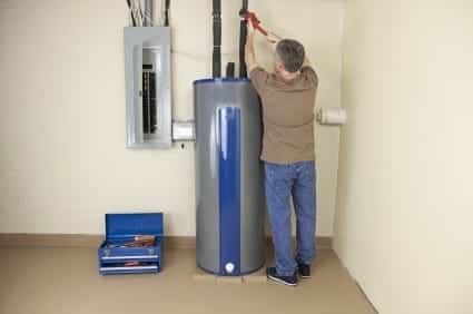 A person is using a wrench on pipes connected to a tall, cylindrical water heater. A blue toolbox is open on the floor nearby.