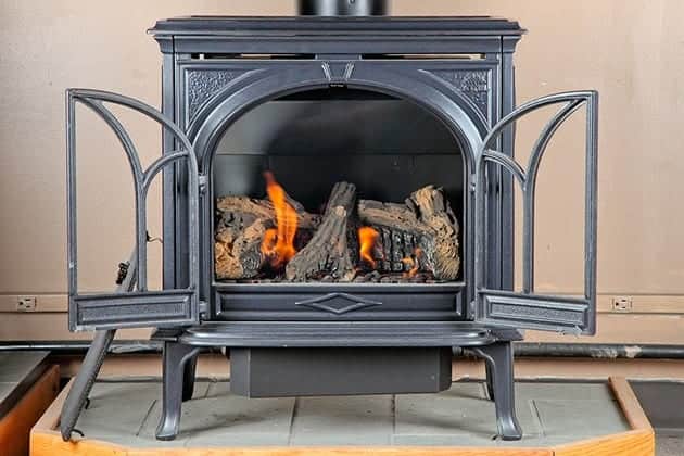 A black cast iron wood stove with open glass doors displays a lit fire with realistic logs on a tiled base.