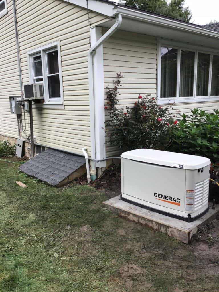 Outdoor generator installed near a house with beige siding, beside a garden with flowers and shrubs. A basement window and an air conditioning unit are visible.