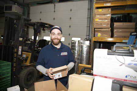 Man in a warehouse smiling, holding a small box. Wearing a blue cap and shirt. Forklift and shelving with boxes in the background.