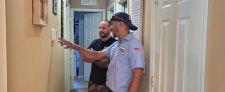 Two men are standing in a hallway. One, in a uniform and cap, points at a thermostat. The other, wearing a black shirt, listens attentively.